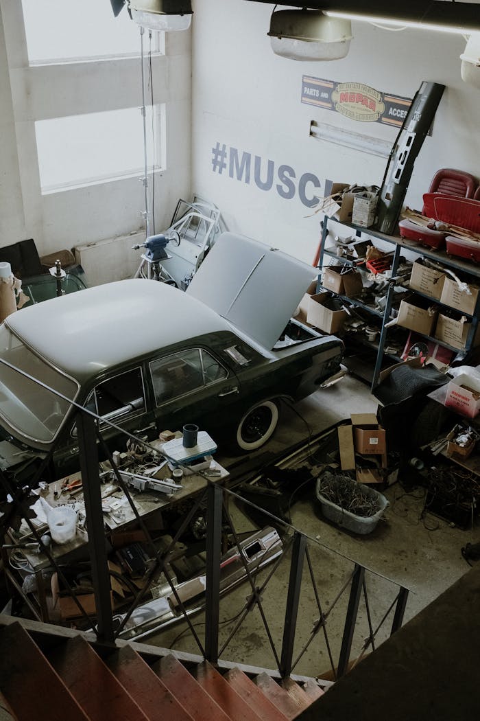 A vintage car is being repaired in a cluttered automotive workshop with tools and equipment.